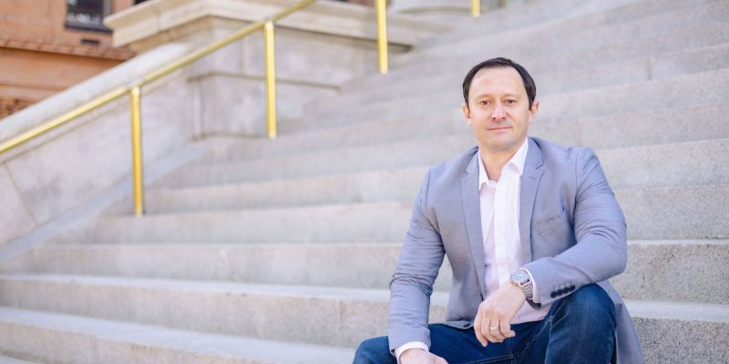 Man in a gray blazer sitting on stone steps outside a building with columns in the background.
