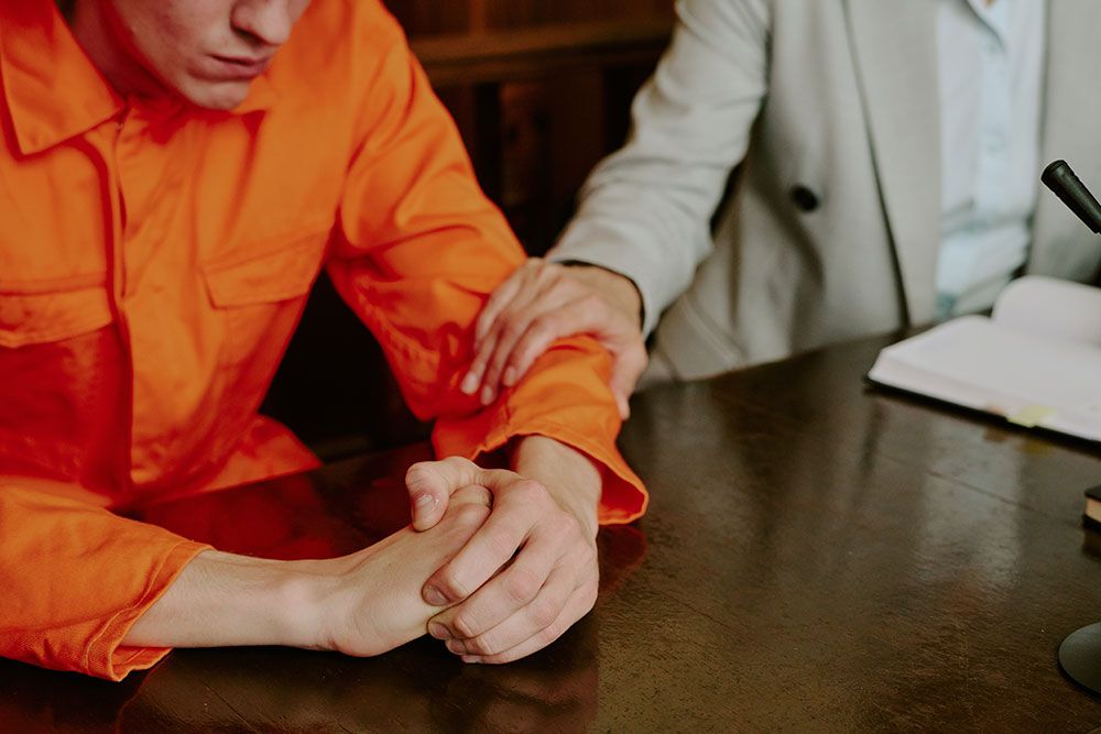 Person in an orange prison jumpsuit sits at a wooden table, hands clasped, with a suited person resting a hand on their arm.