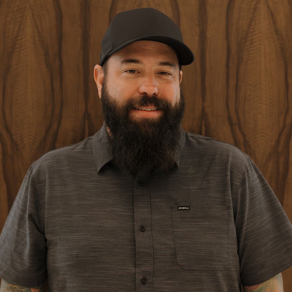 Man with a beard wearing a black cap and gray shirt, standing against a wooden background, smiling slightly.